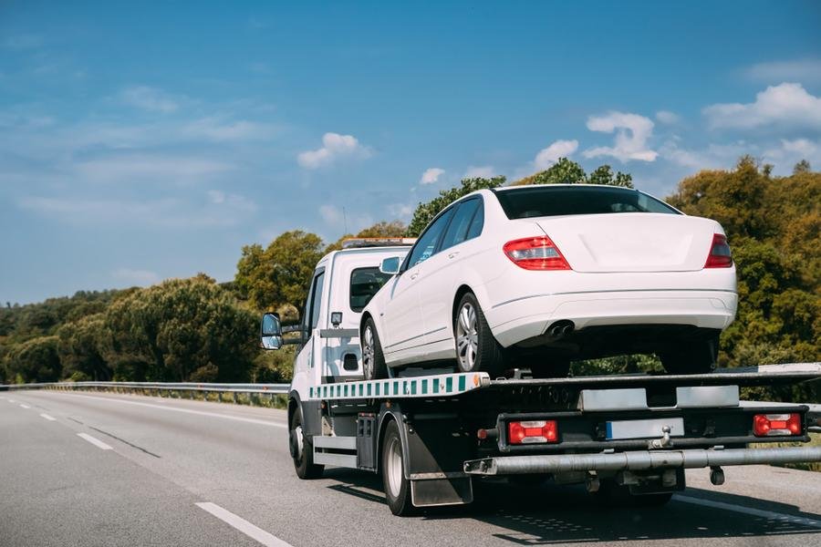 White sedan on a flatbed tow truck being taken to a mechanic shop on the highway